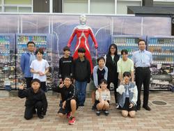 自動販売機が並ぶ建物の前で、赤と銀のウルトラマン像を背景に、先生と生徒たちが集まりピースサインなどをして写した記念写真