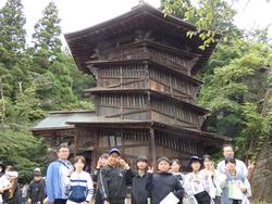 木々に囲まれた中にそびえ立つ、木造三層構造の古い建物の前に並んだ生徒たちがピースサインをして写した記念写真