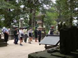 木に囲まれ、大きな石灯籠などが立つ広場で、生徒たちが石碑の方を向いて立っている写真