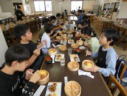 横一列に並んだ食堂のテーブルに対面で座った生徒たちが、楕円形の容器に入った弁当を楽しんでいる様子の写真