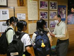 たくさんの古い写真や資料が掲示された壁の前で、3人の女子生徒が男性の説明を聞いている様子の写真