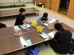 数台の座卓が並べられた広い部屋で、1か所に集まり床に座った3人の男子生徒が、黄色の水入れや資料が置かれた机を囲み、絵付けをしている様子の写真