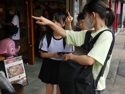 店が並んだ通りに立った3人の女子生徒が、資料を確認しながら行き先を指さし、相談をしている様子の写真