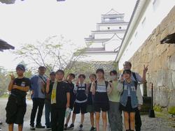 石垣の上に立つ白いお城と木を背景に、城壁の横に並んだ生徒たちと先生がピースサインをして写した記念写真