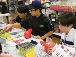 土産物が並んだ店の一角で、新聞紙を敷いたテーブルに横並びに座った3人の男子生徒たちが、それぞれ赤べこに絵付けをしている様子の写真