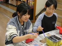 新聞紙が敷かれたテーブルに横並びに座った2人の女子生徒が、赤べこに絵付けをしている様子の写真