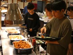 テーブルに様々な料理が並んだビュッフェ形式のレストランで、器を乗せたトレイを持った複数の生徒たちが、順番に料理をよそっている様子の写真