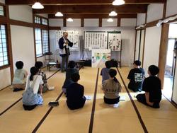 広い和室に前後2列に並んで座った8人の生徒たちが、前方の壁に並べた資料の前に立った、法被姿の男性の話を聞いている様子の写真