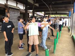 白壁の木造の建物の屋根の下で、緑色のポールの前に立った生徒たちが弓矢を引いて弓矢体験を行っている様子の写真