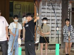 複数の女子生徒が後方で見守る中、1人の男子生徒が弓矢を引いて狙いを定めている弓矢体験の様子の写真