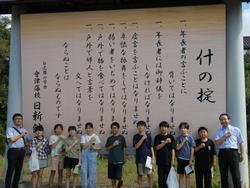 「什の掟」が記された大きな看板の前に並んだ9人の生徒が、両端に立った男性の先生2人と一緒に左胸に拳を当てたポーズをとって写した記念写真