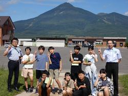 複数並んだ建物の後ろにそびえる緑の山を背景に、9人の生徒と2人の男性の先生が並んでピースサインをして写した記念写真