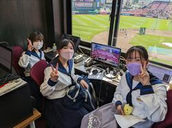 野球場の放送席に座った3人の女子生徒がピースサインをしている写真