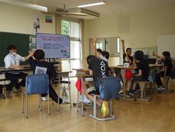 役場税務課の職員の方が椅子に座って横にあるモニターを指さし、複数の生徒が手を挙げている租税教室の様子の写真