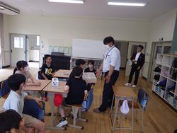 教室で、複数の生徒たちが机を囲んで座り、レプリカの1億円を見ている様子の写真