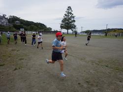 曇り空の下、グラウンドで帽子をかぶった小学生たちが、各自のペースで持久走をしている様子の写真
