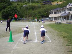 体操服を着用した2人の子供が、スタートラインに立ち、赤い旗を持った男性の合図で走り出そうとしている様子の写真