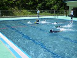 屋外プールで5人の生徒たちが、顔を水につけバタ足で泳いでいる写真