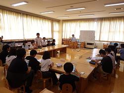 明るい室内で、複数の学生たちが机を囲んで座り、そのうちの一人が皆の前で発表をしている、代表委員会の様子の写真