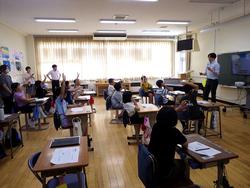 黒板の前で男性が話をしており、生徒たちが一斉に手を挙げている、活気あふれる出前授業の様子の写真