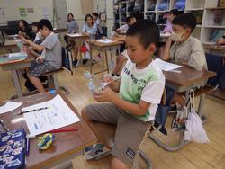 ペットボトルを使い、熱心に工作に取り組んでいる小学生と、机に向かって座っているクラスメイトを写した教室の写真