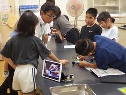 黒いテーブルで、先生が複数の生徒に何かを教えたり、生徒がタブレット端末を見ながら実験器具のようなものを組み立てたりしている様子の写真