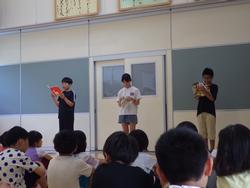 広い室内で、三人の小学生が前に立って朗読し、それを床に座った大勢の児童たちが静かに聞いている様子を写した写真