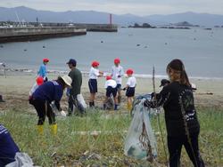 防波堤のある海岸で、赤白帽をかぶった小学生たちが先生たちと一緒に、トングとごみ袋を持って海岸のごみ拾いをしている様子の写真