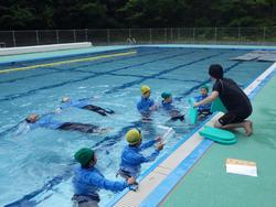屋外のプールで、複数の子どもたちが服を着用して泳ぎの練習をしており、先生が指導している様子の写真