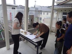 受付の机で書類に記載している保護者と、近くには引渡しを待つ子どもたちや学校関係者の方々が写っている引渡訓練の様子の写真