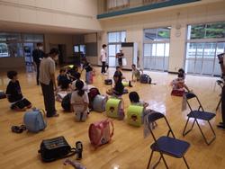 ランドセルや荷物を持って室内に集まり、保護者の迎えを待っている生徒たちの様子を写した写真