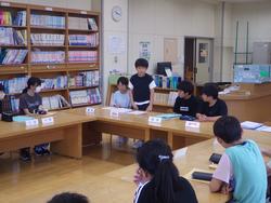 図書室にコの字に設置された机に代表委員の生徒たちが席について話し合いが行われており、一人の男子生徒が自席に立って、発表している様子の写真