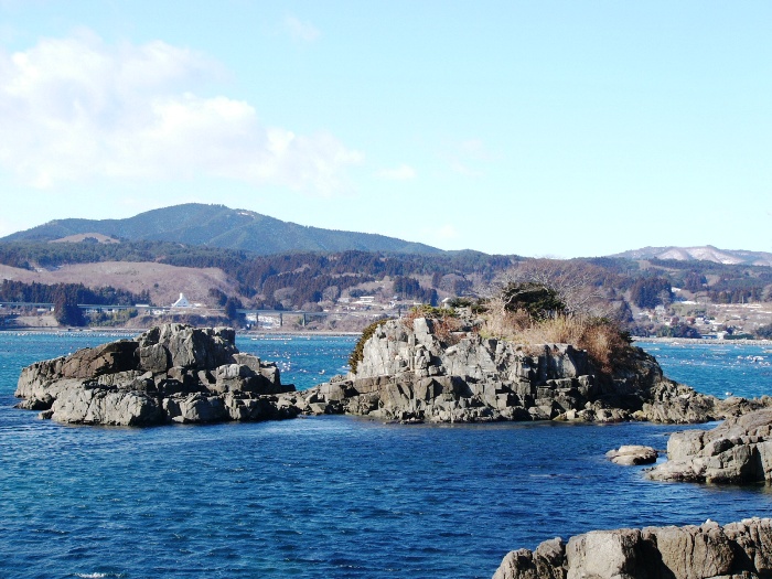 手前にゴツゴツとした岩場、奥に家々が見える入り江と青い海が広がる写真