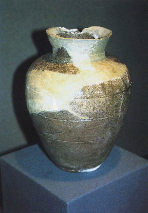 欠けや修復跡のある茶褐色と灰色がかった陶器の壺「三筋壷」が展示台に置かれている写真