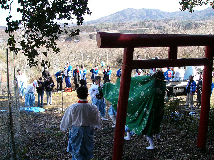 赤い鳥居をくぐった先で獅子舞が演じられ、観客が集う山里の祭りの写真