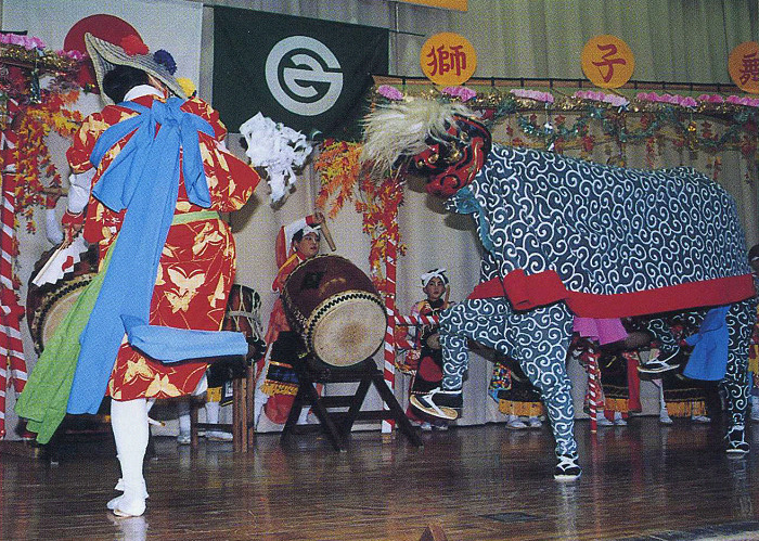 花飾りなどで彩られた舞台で、赤地に蝶模様の着物を着た演者と、獅子舞の演者が舞う様子の写真