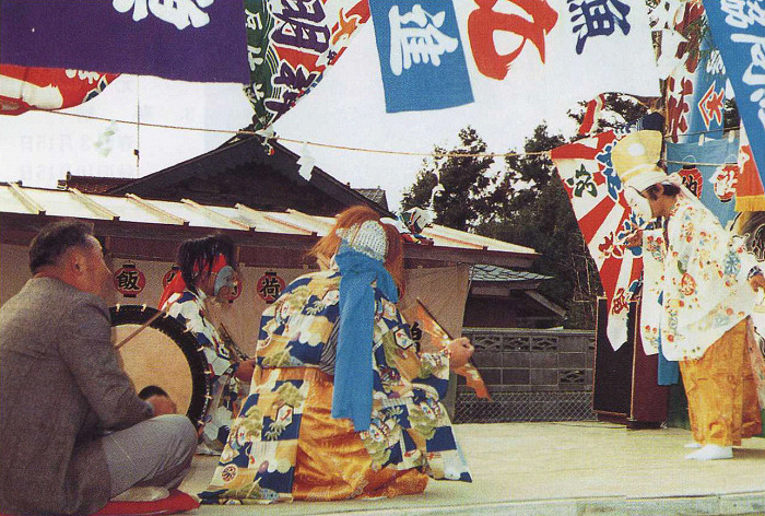 色鮮やかな着物と獅子舞の面を着けた人々が太鼓の伴奏で舞台上で伝統的な舞を披露し、背後には多彩な旗や神社の屋根が見える屋外写真