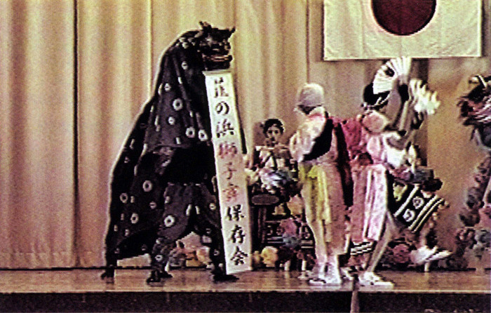 舞台上で獅子舞が「韮の浜獅子舞保存会」と書かれた札を持ち、色鮮やかな衣装の人物とともに日本国旗の下で演舞する様子の写真