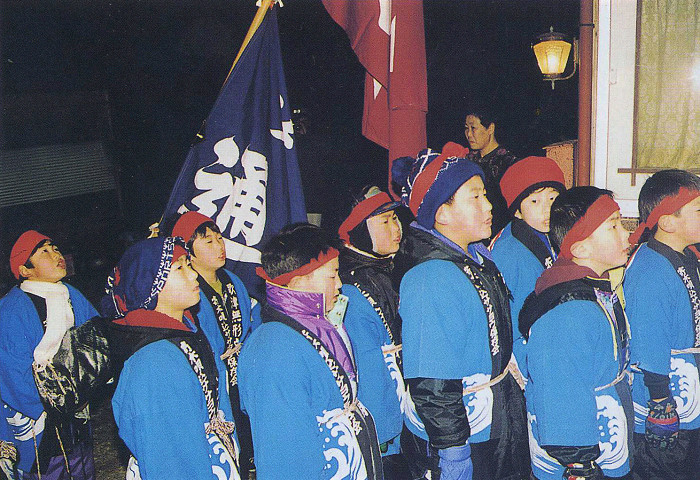 青い法被と赤い鉢巻を身につけた子どもたちが夜の屋外で整列し、後方に旗を掲げながら一方向を見上げている様子の写真