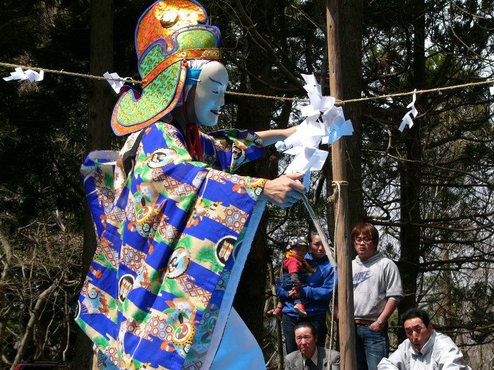 色鮮やかな青地の装束と大きな飾り帽子、白い能面を着けた人物が屋外のしめ縄の下で舞いを披露し、周囲の人々が見守る様子の写真