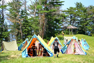 青とオレンジの三角テントがいくつも立てられており、多くの子どもたちがテントの間で活動している夏のキャンプ場の写真