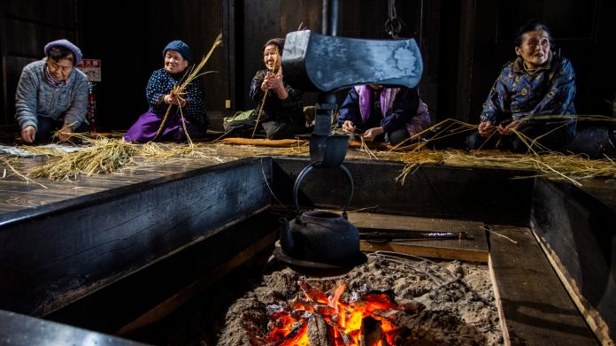 囲炉裏の周りに数人が座り、手作業で藁を編んでいる様子が写っている写真