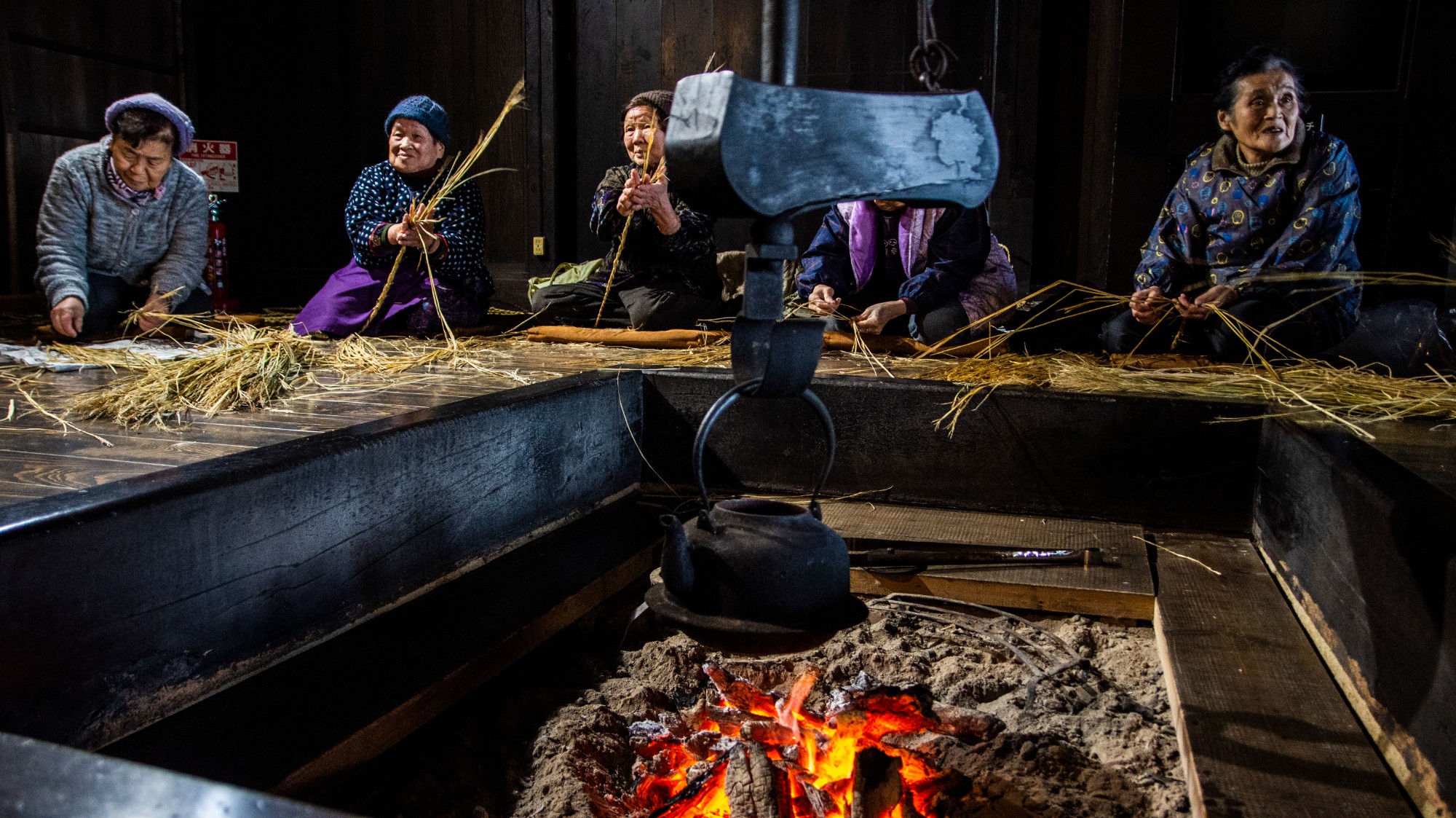 囲炉裏の周りに5人の年配の女性が座り、手作業で藁を編んでいる様子が写っている写真