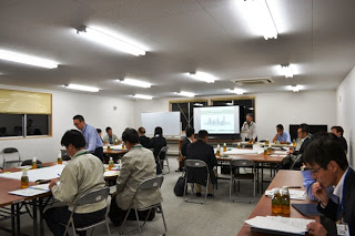 推進会議の参加者がいくつかのグループに分かれて着席しており、机の上の資料が広げられている会議室全体の写真