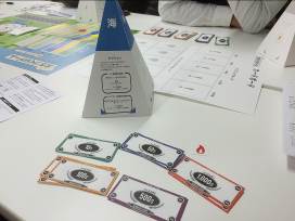 テーブルの上に、「海」と書かれた紙製の三角すい、複数の紙幣風カードが並び、その後ろに「まちづくりゲーム」の用紙などが置かれている写真