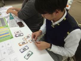 参加者の男性が、テーブルの上に並べられた紙幣風のカードを手に取り、ゲームに取り組んでいる様子の写真