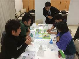 5人の参加者がみんなのまちづくりゲームの用紙を広げたテーブルを囲み、ゲームをしており、傍に立ったスーツ姿の男性がゲームをのぞき込んでいる様子の写真