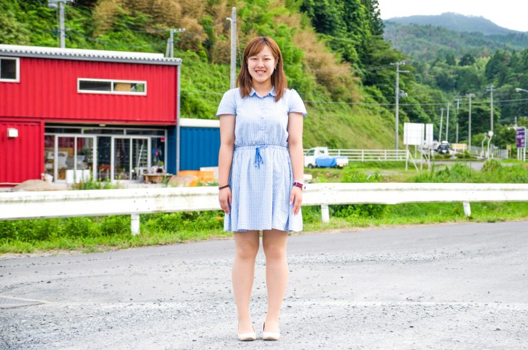 赤と青の外壁の建物の前に立つ、水色のワンピースを着た佐藤さんの写真