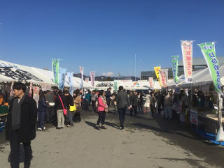 左右にのぼりを立てた白い屋根のテントが立ち並び、たくさんの来場者で賑わう福興市の会場の写真