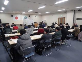 国旗が掲げられた壁の前にホワイトボードが置かれ、たくさん並んだ長机の席に、多くの出席者たちが座っている会場全体の写真